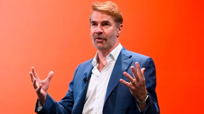 আমাদের কি জেনারেটিভ এআই দরকার? | BanglaKagaj.in Erik Brynjolfsson gestures while speaking onstage during a panel, with a vivid orange background behind him.